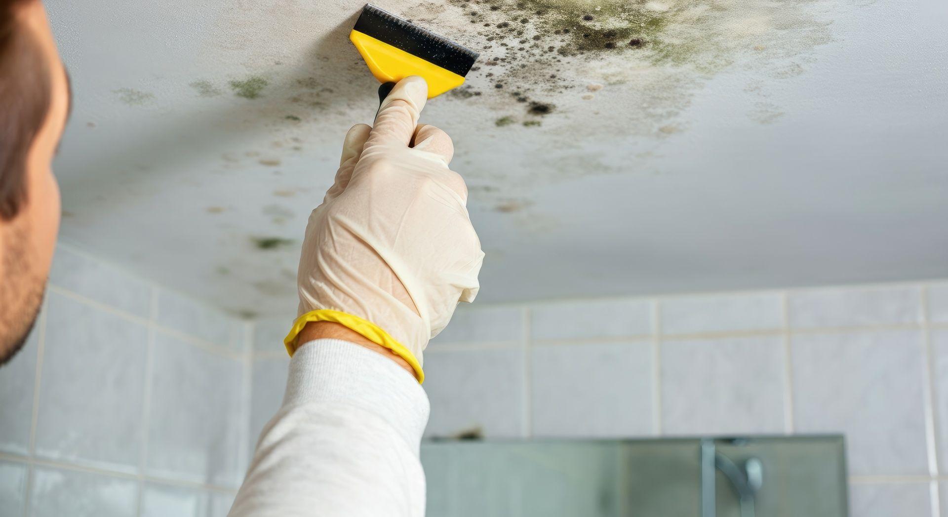 Gloved hand scrubbing mold on a bathroom ceiling with a yellow sponge brush