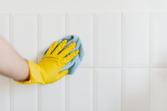 Yellow-gloved hand cleaning white tiled wall with a blue sponge
