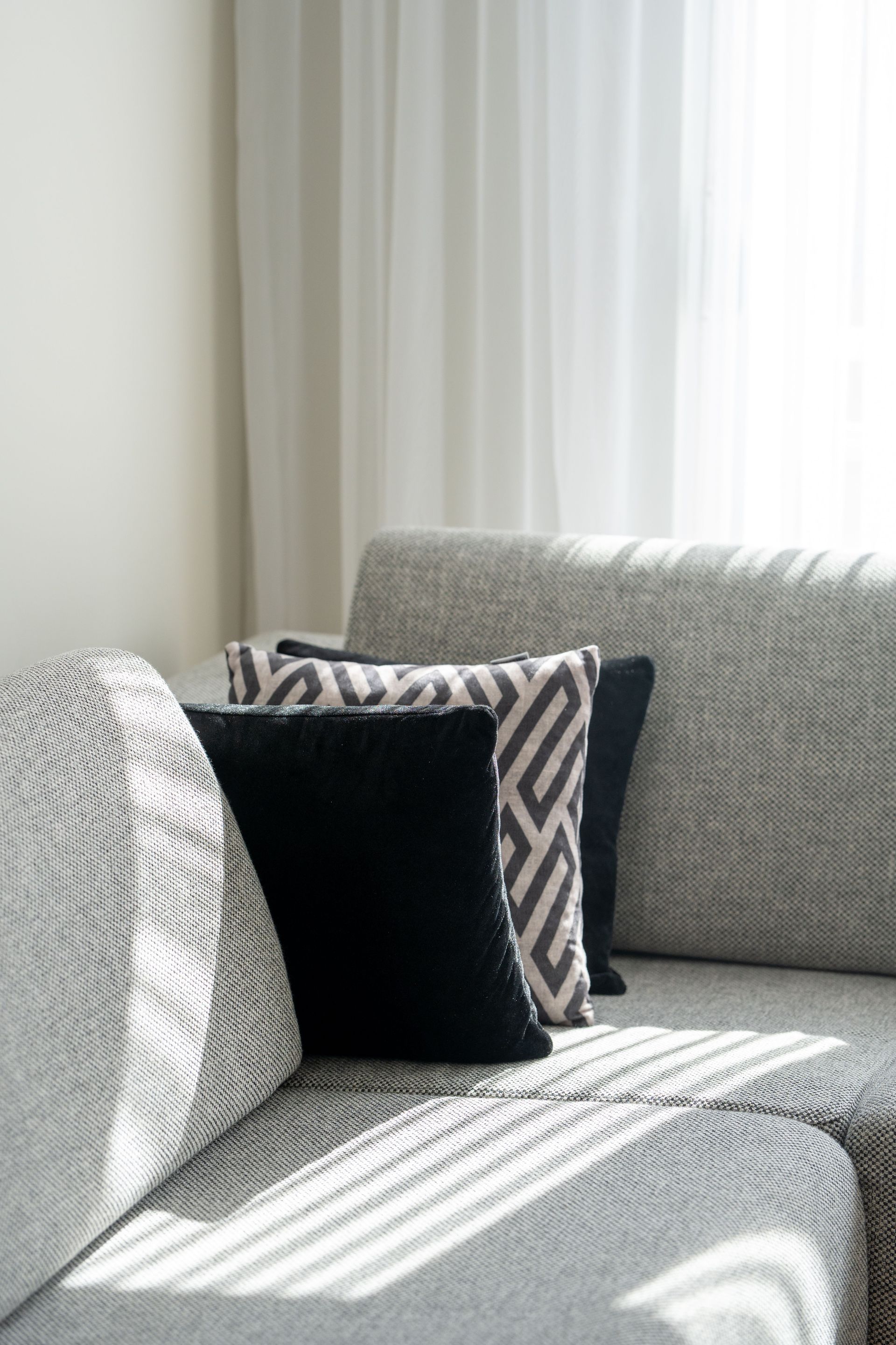 Sunlit sofa with black and patterned cushions beside sheer curtains