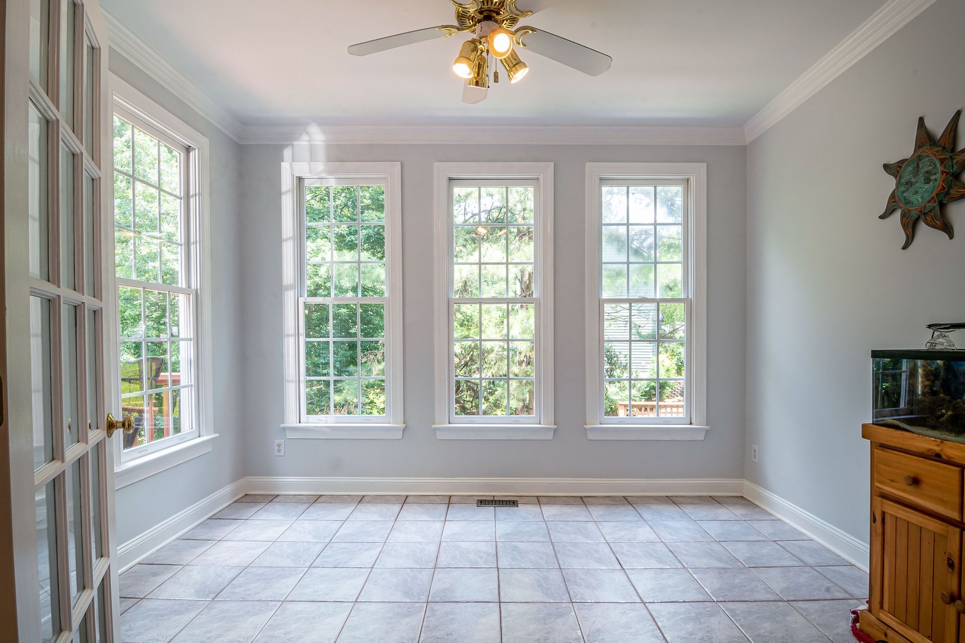 Empty light gray room with tall windows, tiled floor, ceiling fan, and wood cabinet