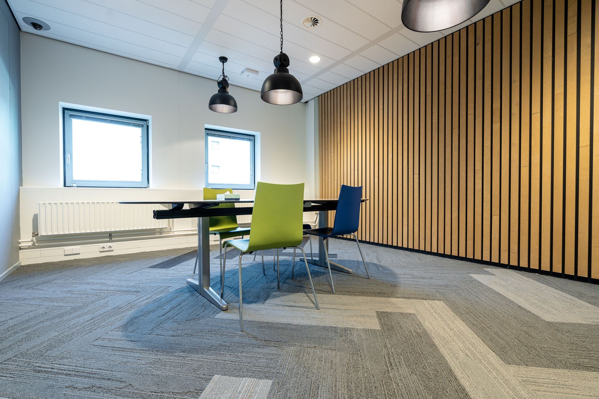 Modern meeting room with round table, green and blue chairs, pendant lights, and wood slat wall.