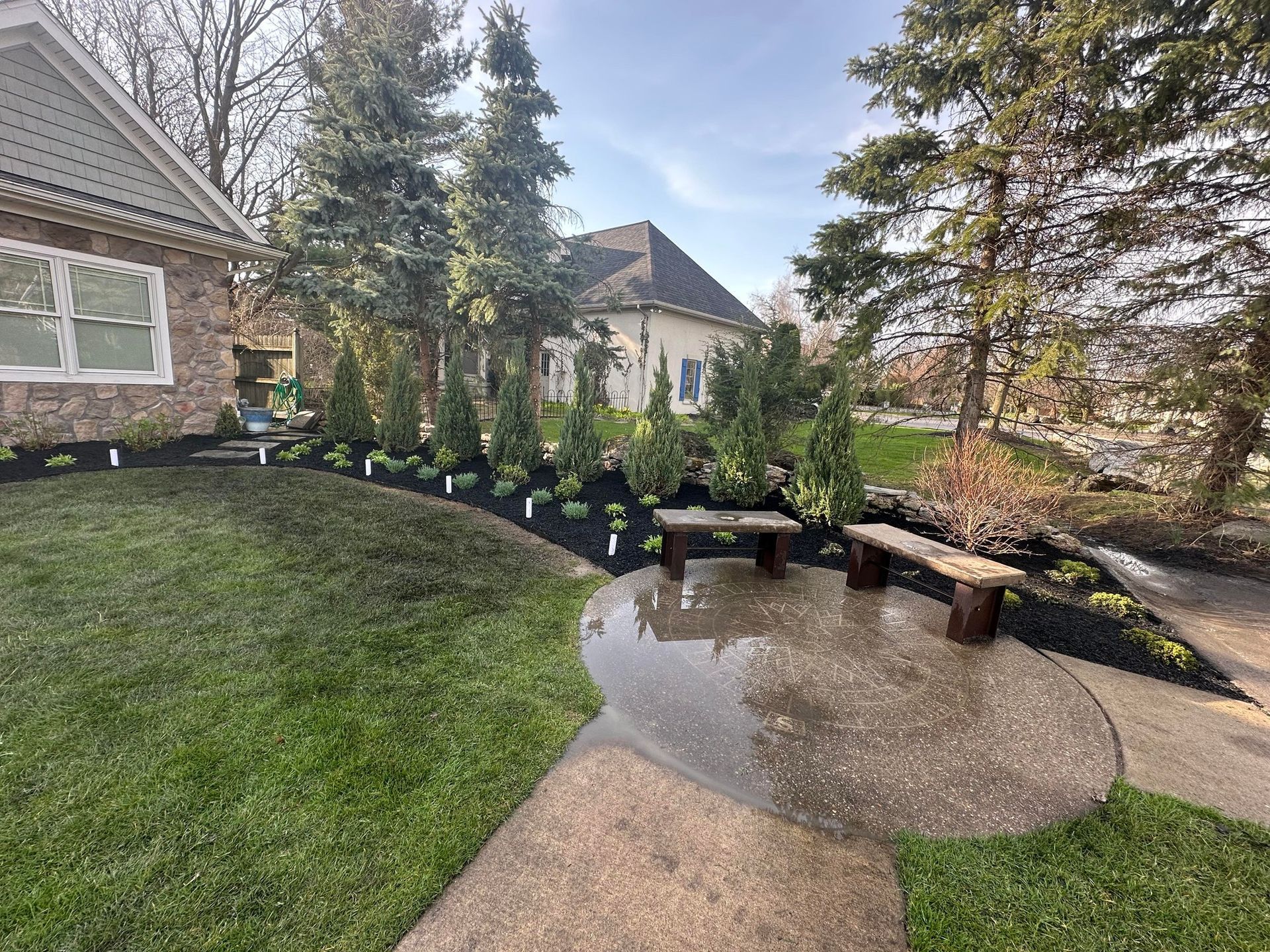 Landscaped front yard with two benches, trees, and dark mulch.