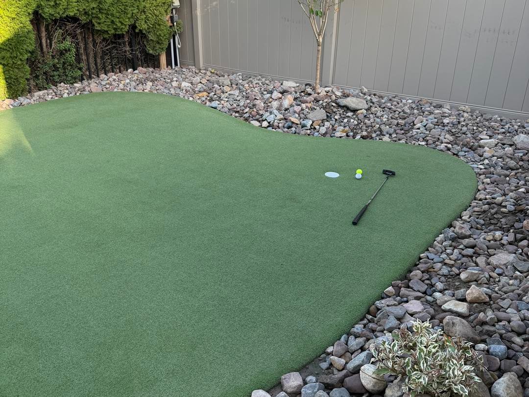Green putting green with golf balls and a putter, bordered by rocks and a fence.