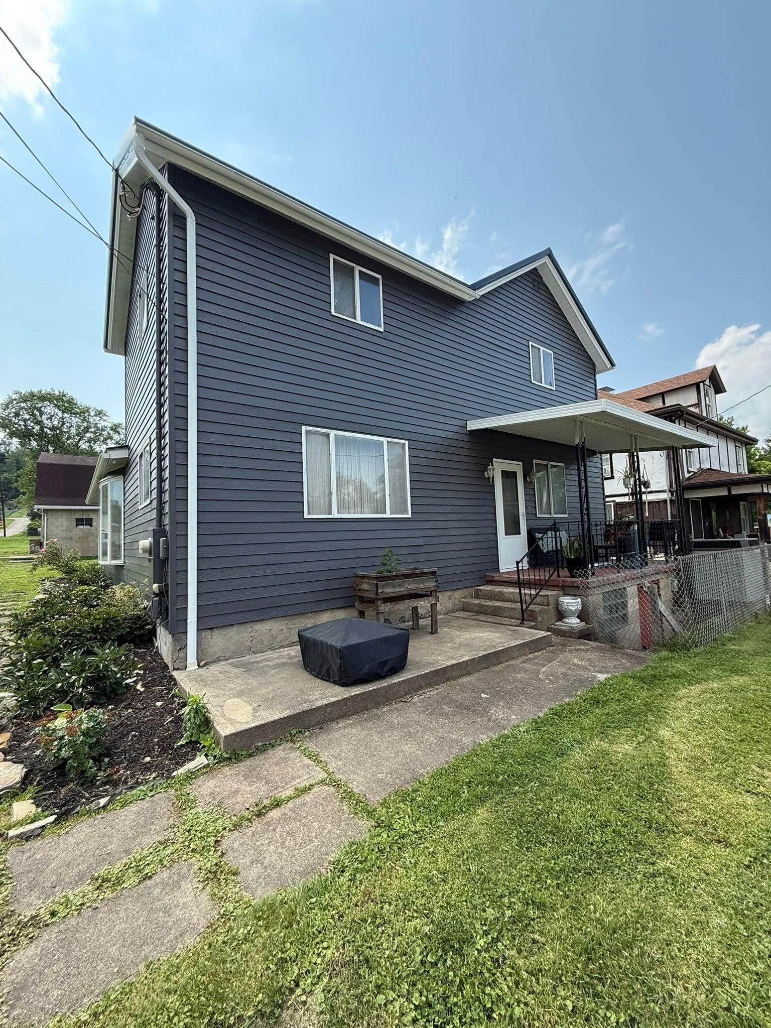 Blue-gray two-story house with a small porch, front garden, and paved walkway under a sunny sky