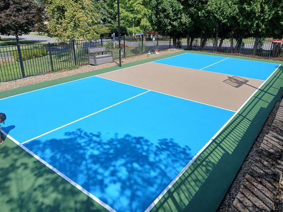 Empty outdoor sports court with blue and tan surface, surrounded by trees and a fence.