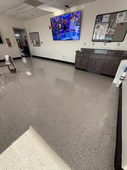 Clinic waiting room with speckled floor, wall-mounted TV, and cabinets along the right wall.