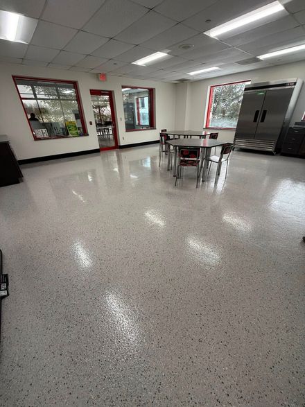 Empty school cafeteria with white tables, red-framed windows, and a speckled tile floor.