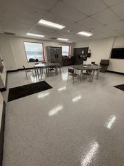 Empty cafeteria with tables and chairs on a glossy speckled floor beneath fluorescent lights.