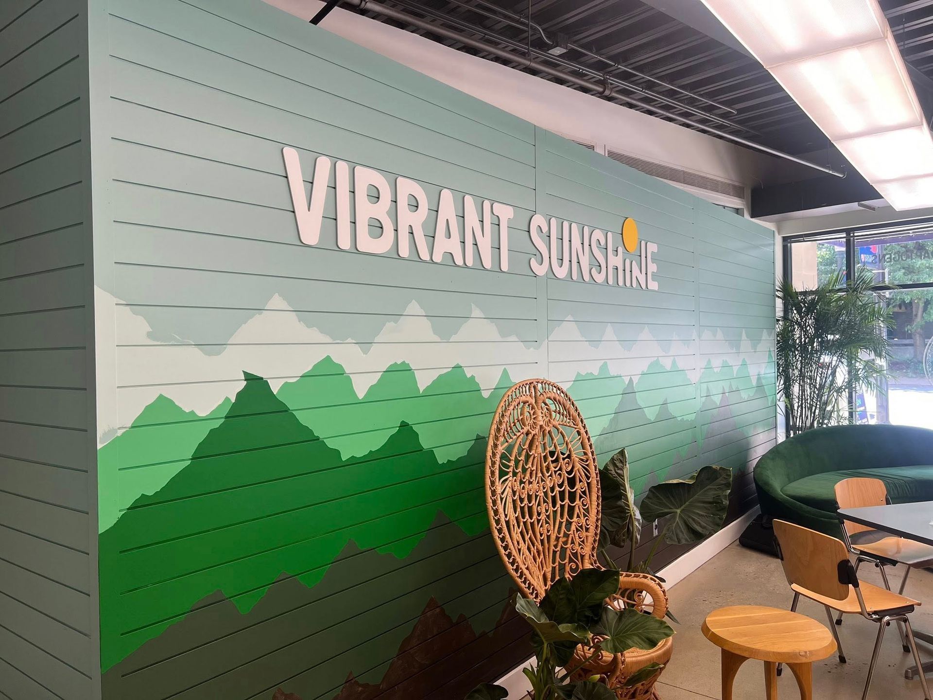 Vibrant Sunshine mural on a wall with green mountains, a wicker chair, and small café tables under a canopy