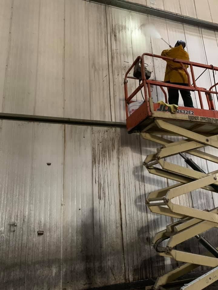 Worker in a yellow jacket on a scissor lift inspecting a tall wall indoors.