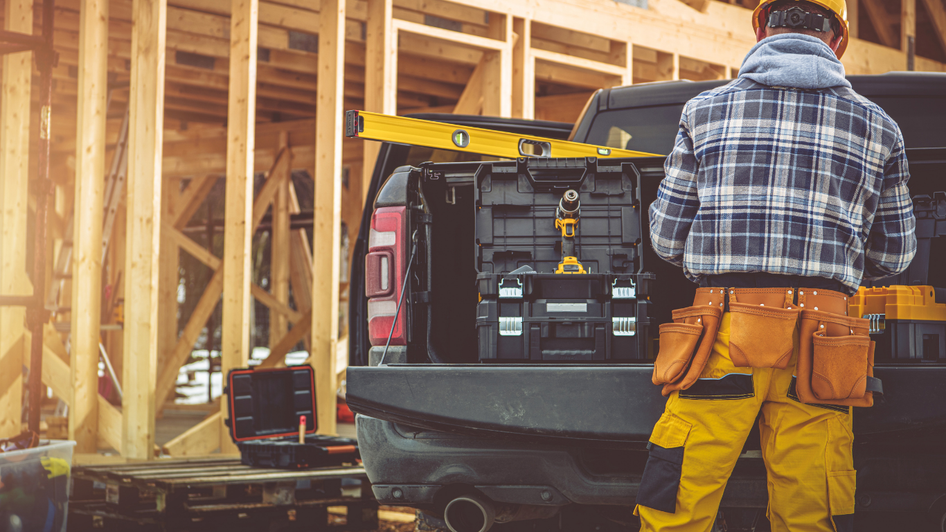 Worker in yellow pants stands beside a truck at a wooden house construction site.