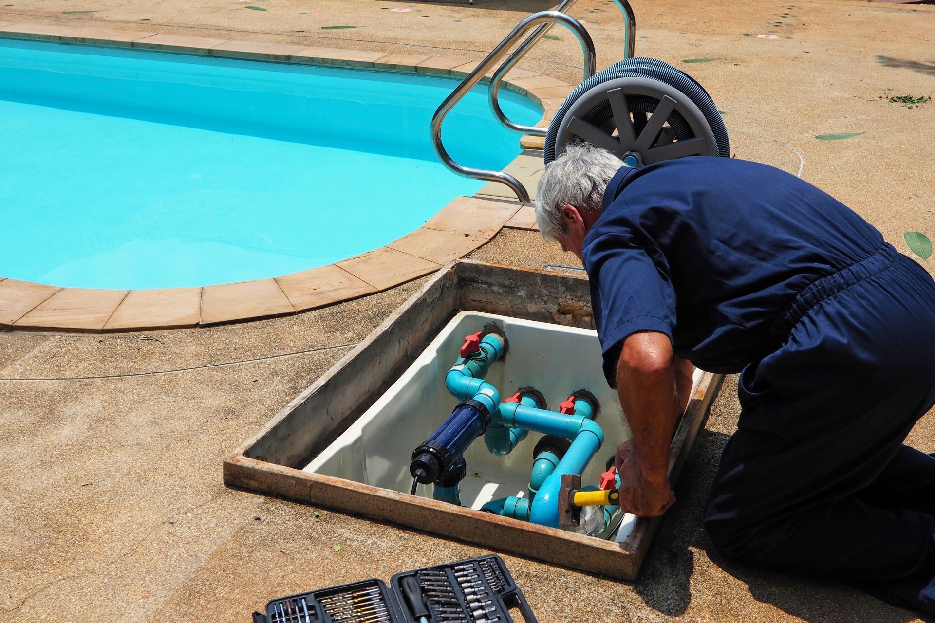 Technician kneeling by poolside pump system, inspecting blue pipes beside a swimming pool