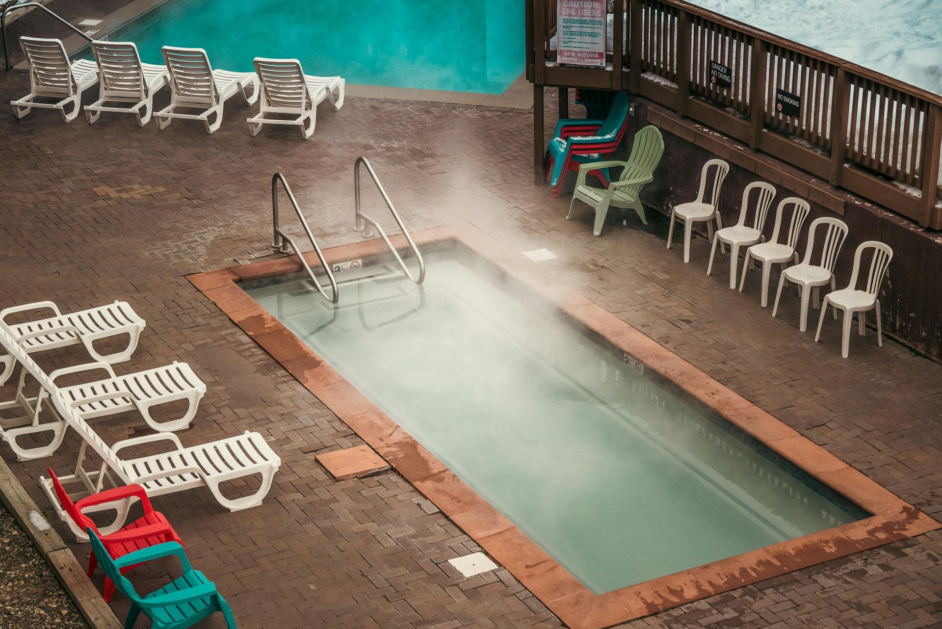 Indoor hot tub with steam, surrounded by lounge chairs and stacked white chairs on a wooden deck