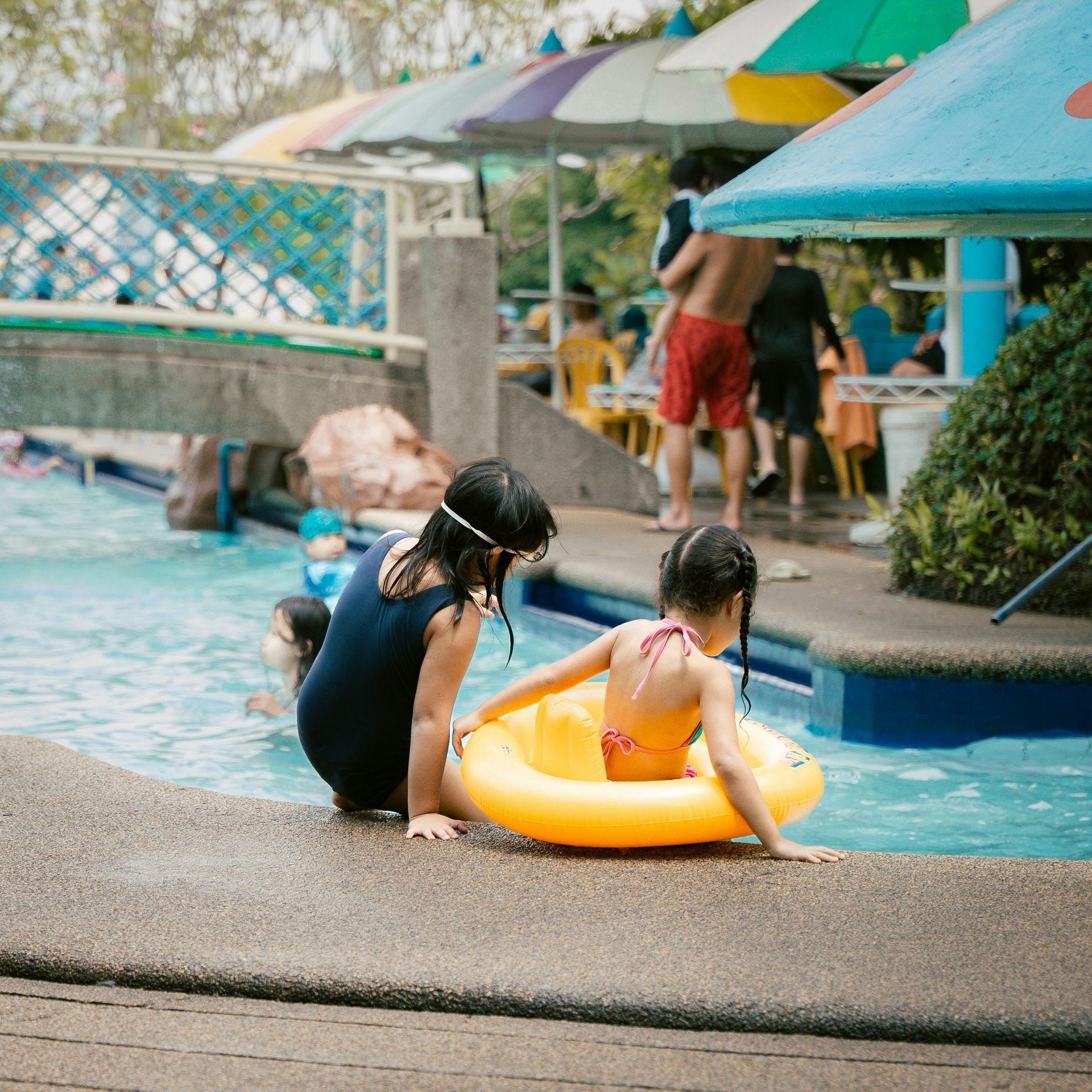 Children playing by a pool, one seated in a yellow float while another stands in the water under umbrellas.