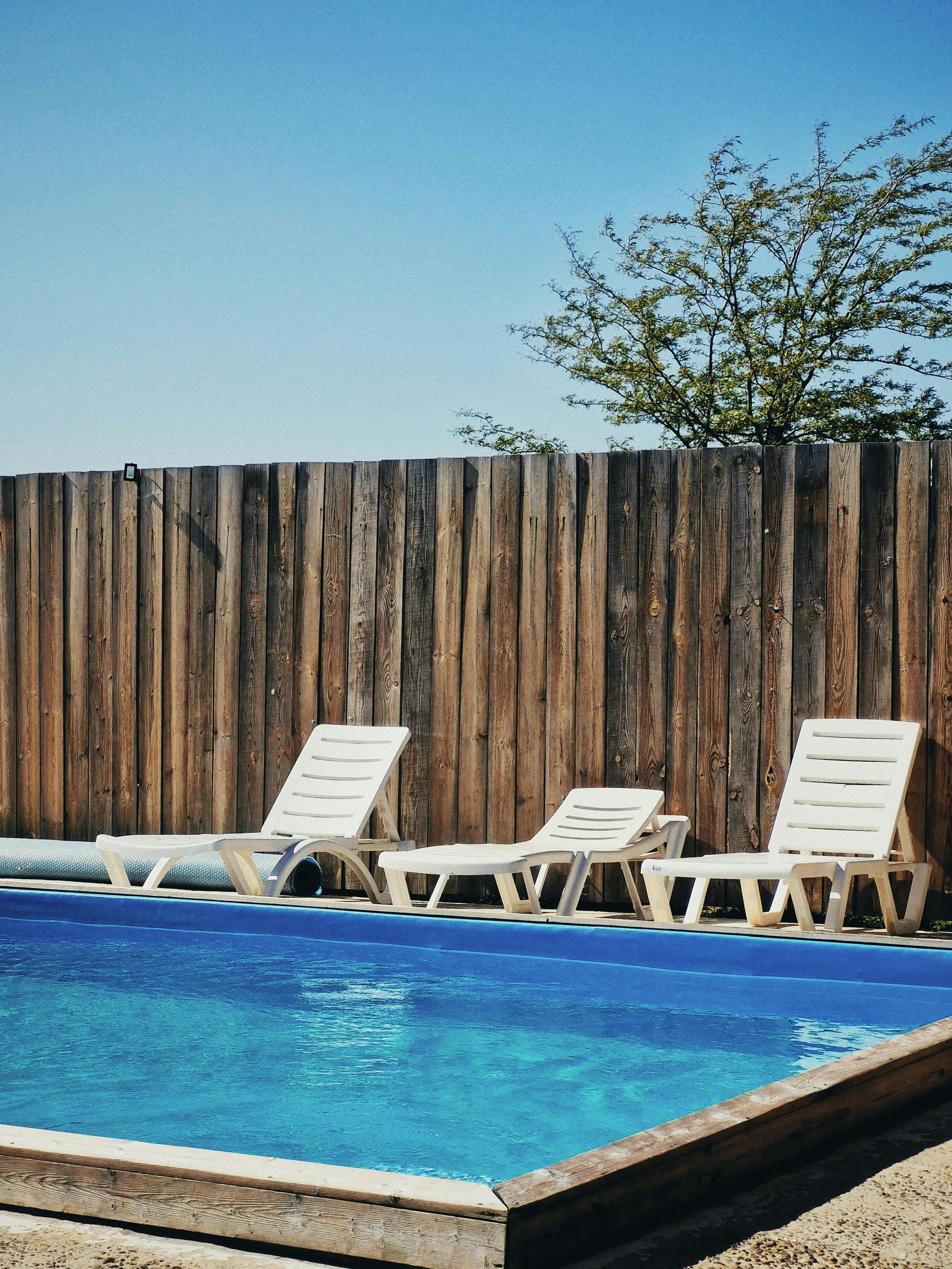 Poolside lounge chairs beside a blue swimming pool and wooden fence under a clear sky