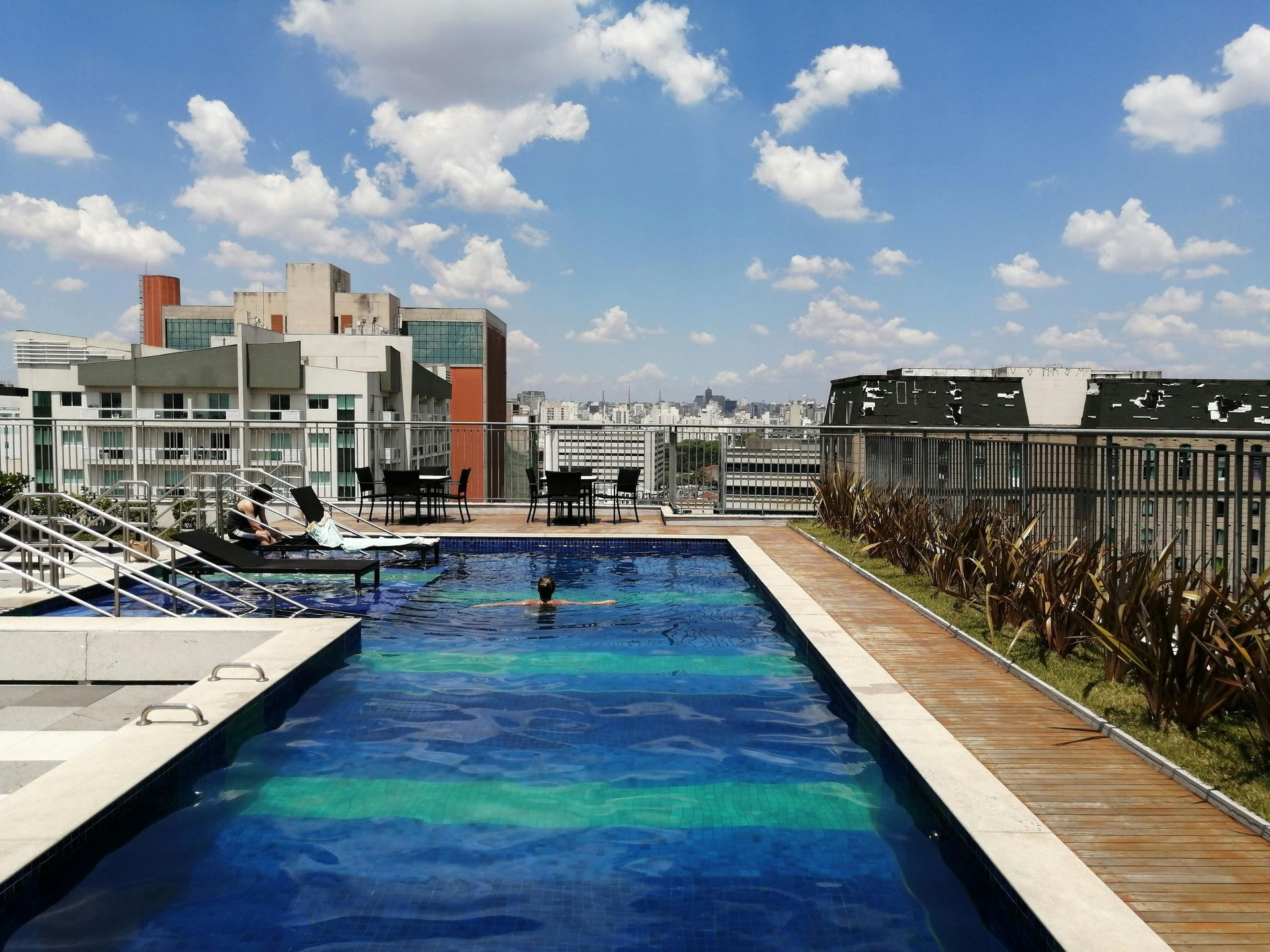 Rooftop infinity pool with blue water, lounge chairs, and city buildings under a partly cloudy sky