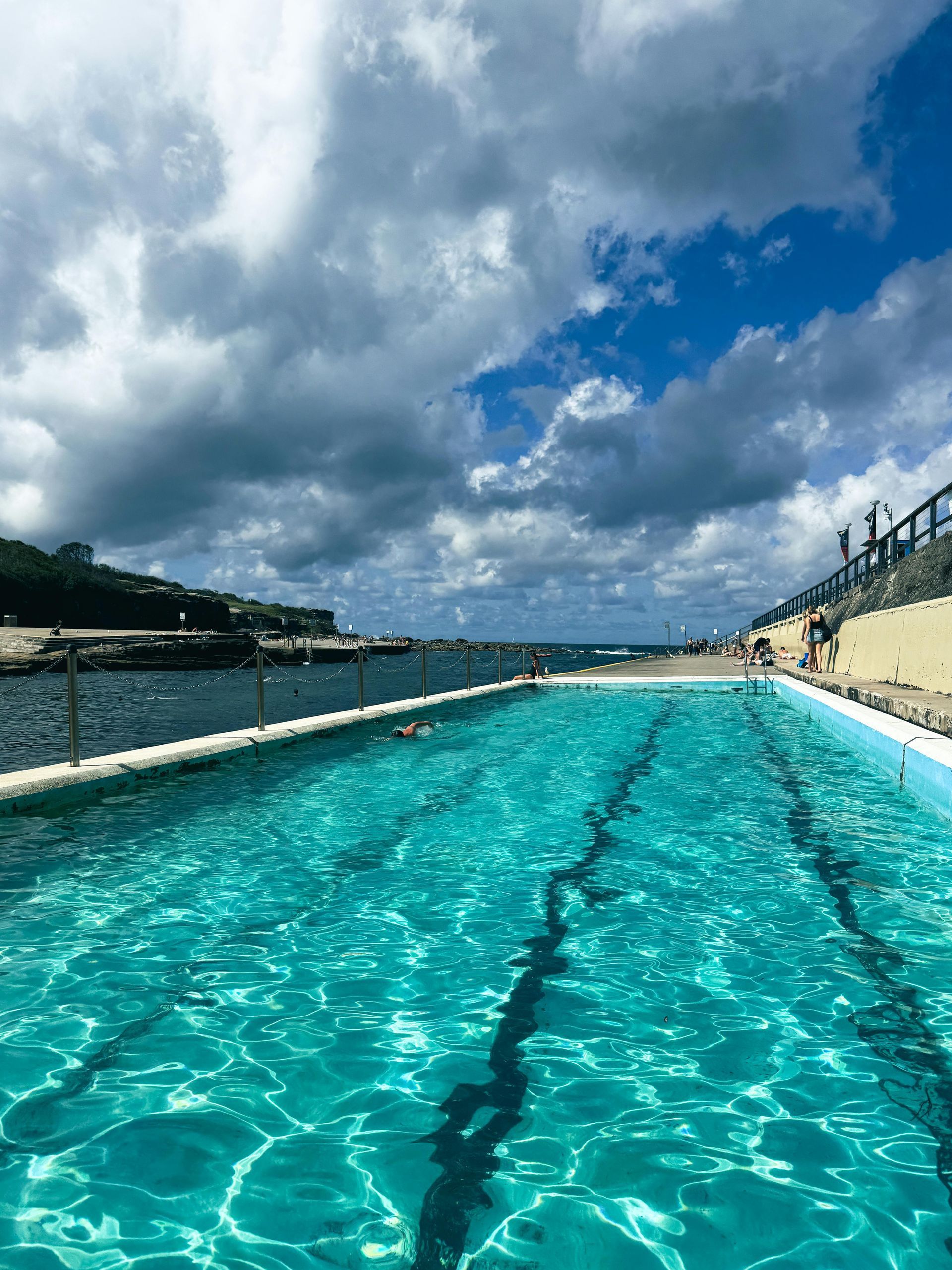 Outdoor lap pool with turquoise water under a cloudy blue sky, beside a sandy beach and hillside seating