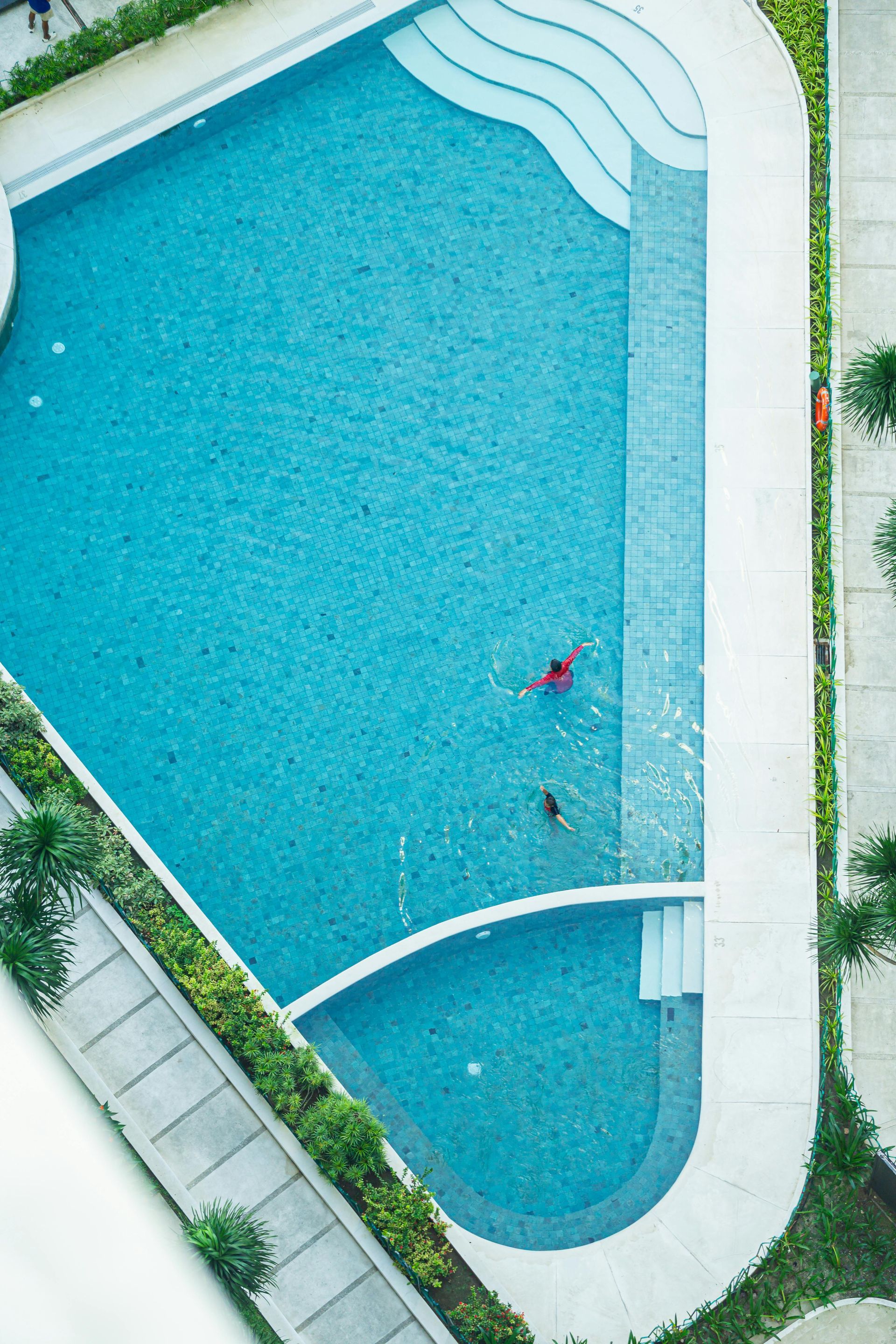 Aerial view of a turquoise swimming pool with curved steps and a small swimmer near the center.