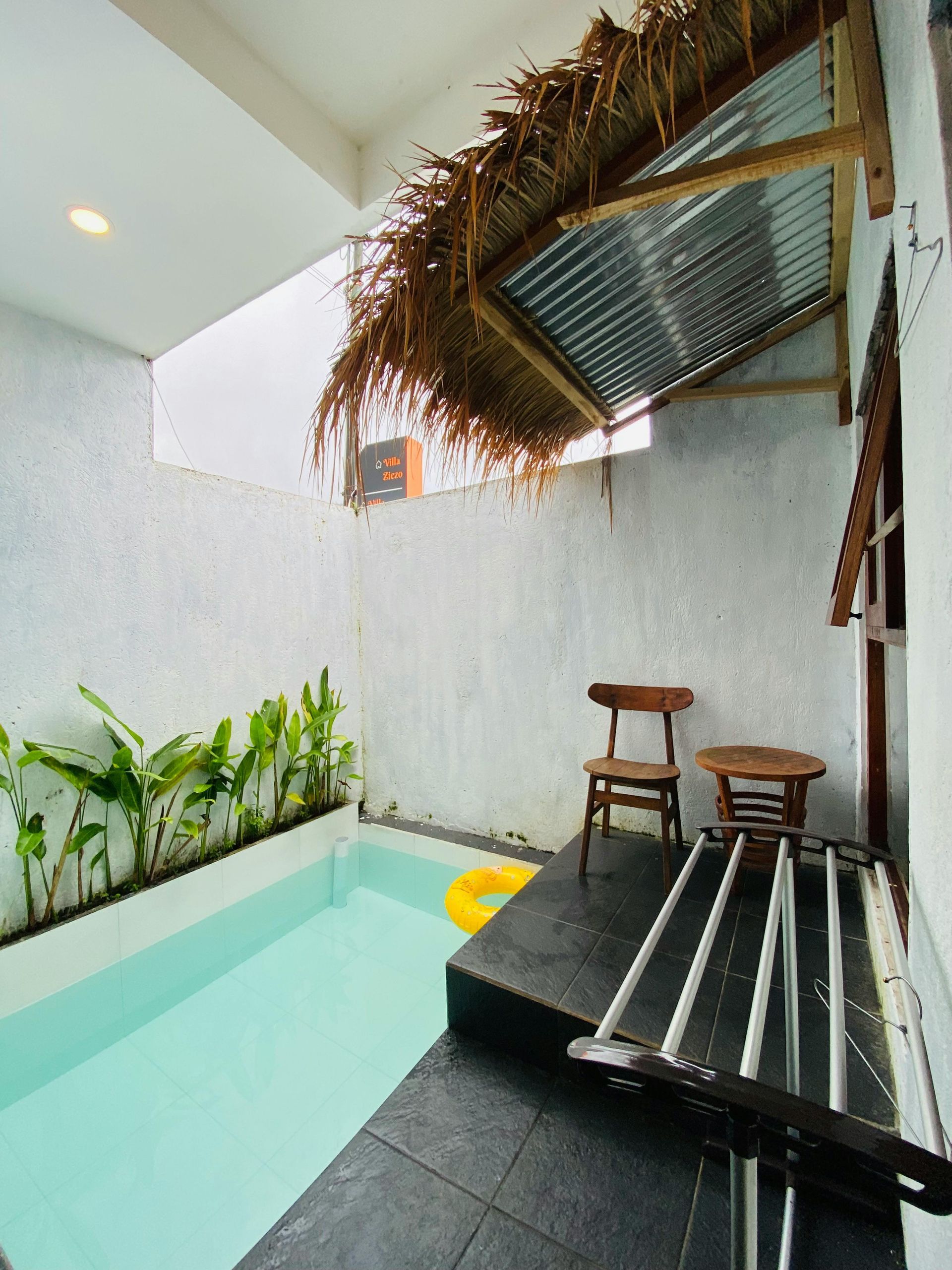 Small tropical courtyard with a narrow pool, black deck, stools, and a thatched canopy
