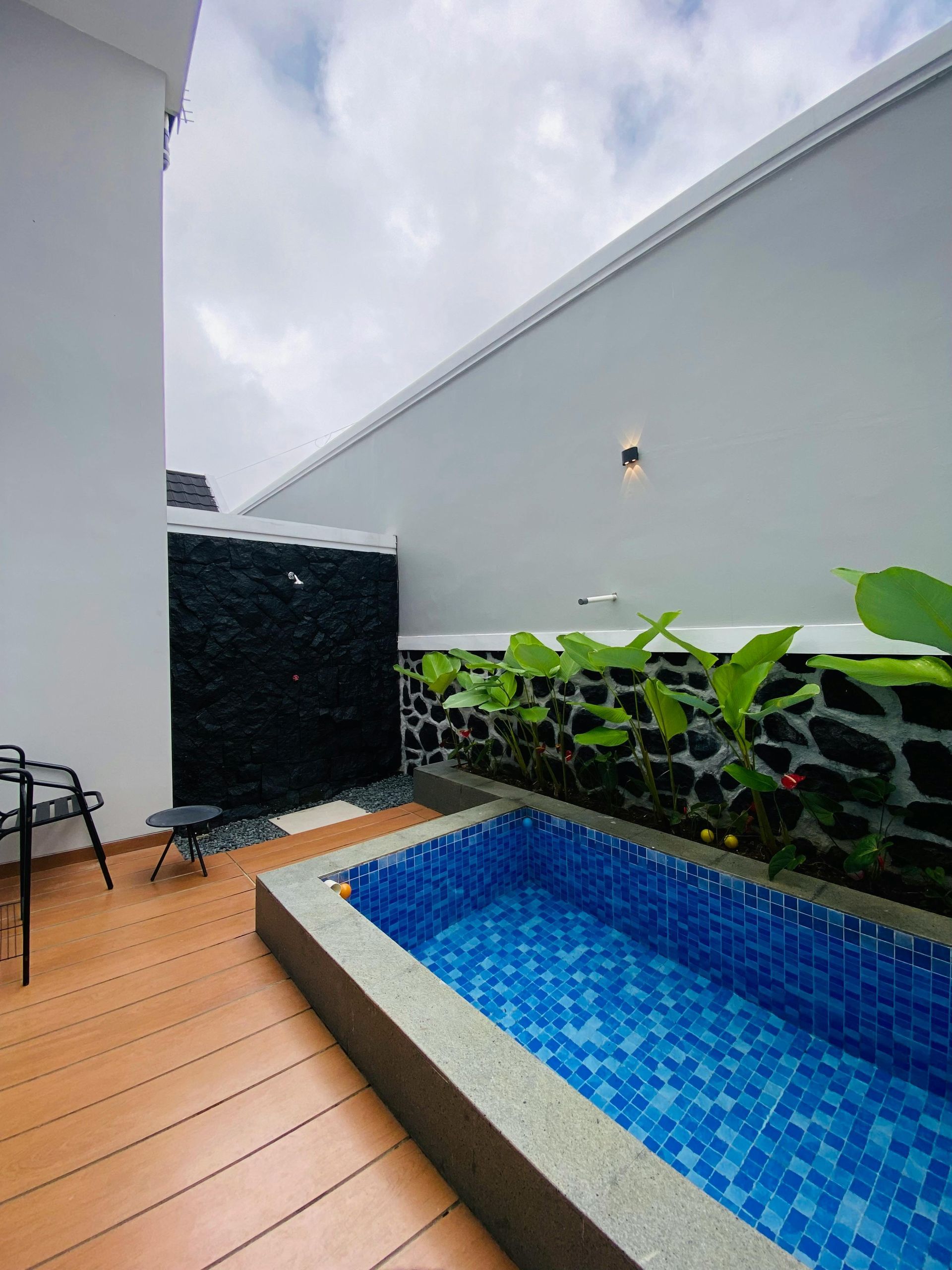 Small patio with a blue-tiled pool, wooden deck, black wall feature, and cloudy sky above.
