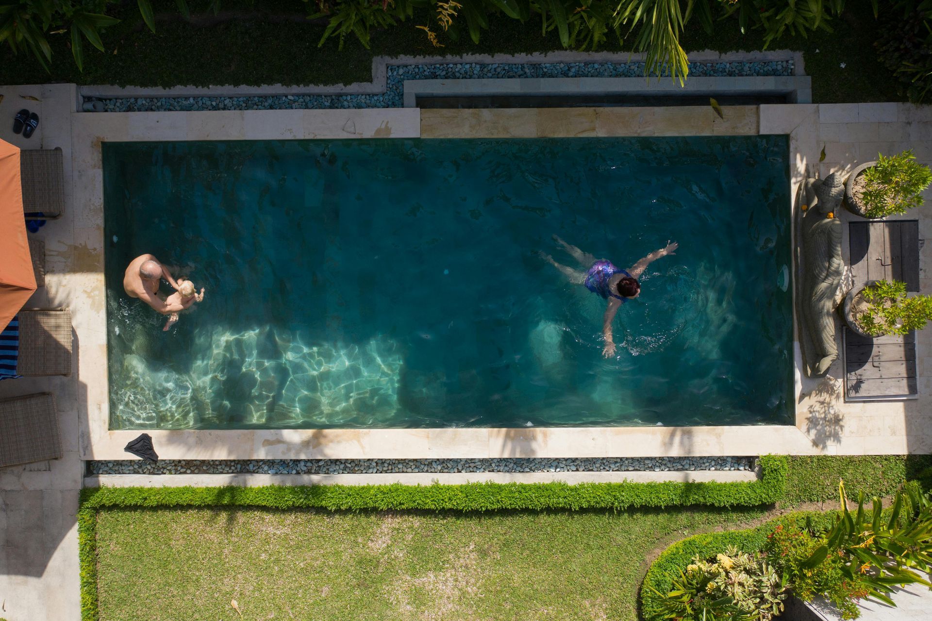 Aerial view of two people swimming in a rectangular backyard pool surrounded by patio and lawn.