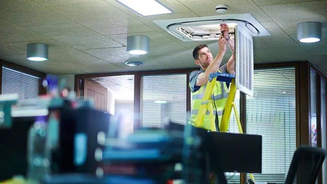 A man on a ladder is installing or repairing an air conditioning unit in an office. He wears a reflective vest.