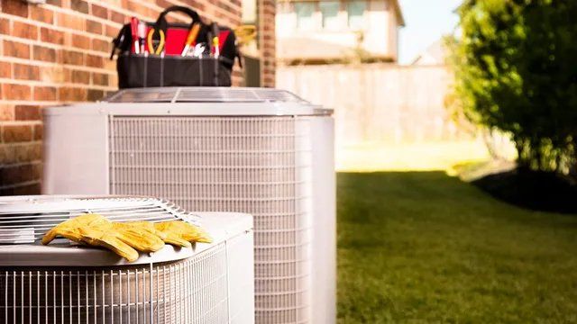 HVAC units outside with a toolbox and yellow gloves on top, in front of a brick wall and grass.