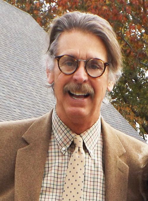 Smiling Pastor Bill in glasses and a tan blazer stands outdoors among autumn trees with the Parish Hall roof behind him