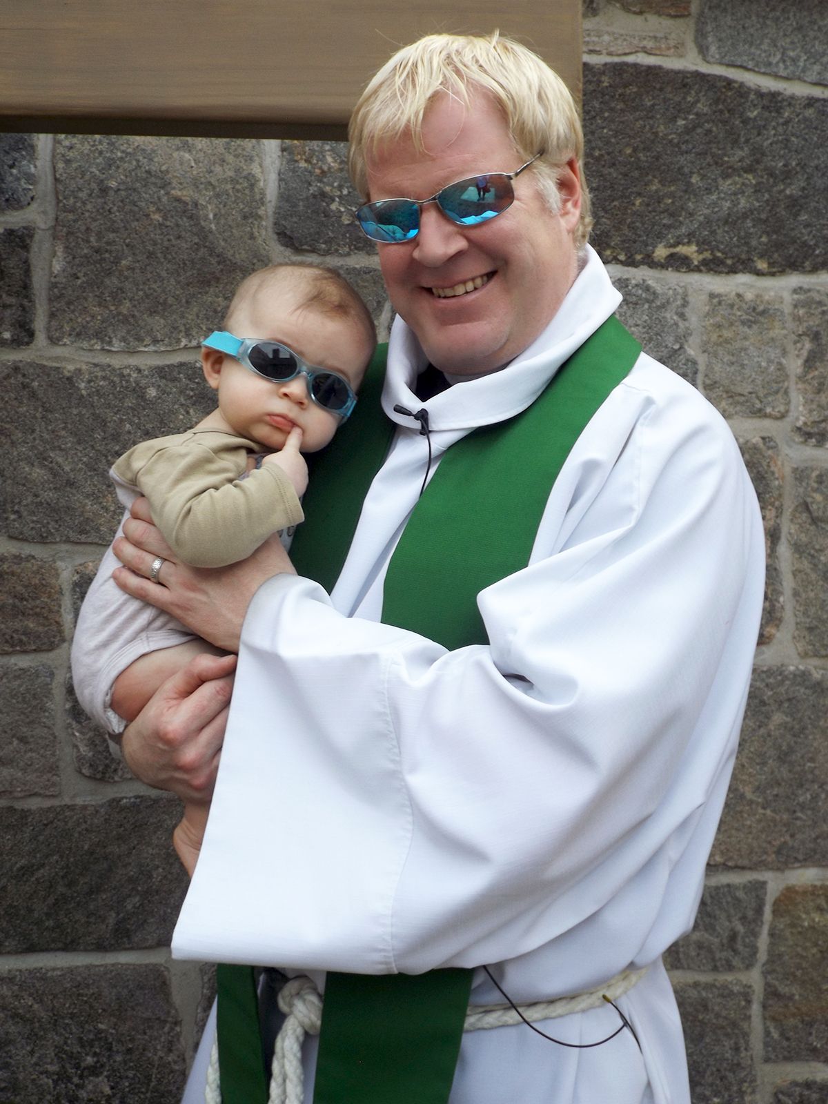 Smiling Pastor holds a baby by a stone wall outdoors, while both wear sunglasses.