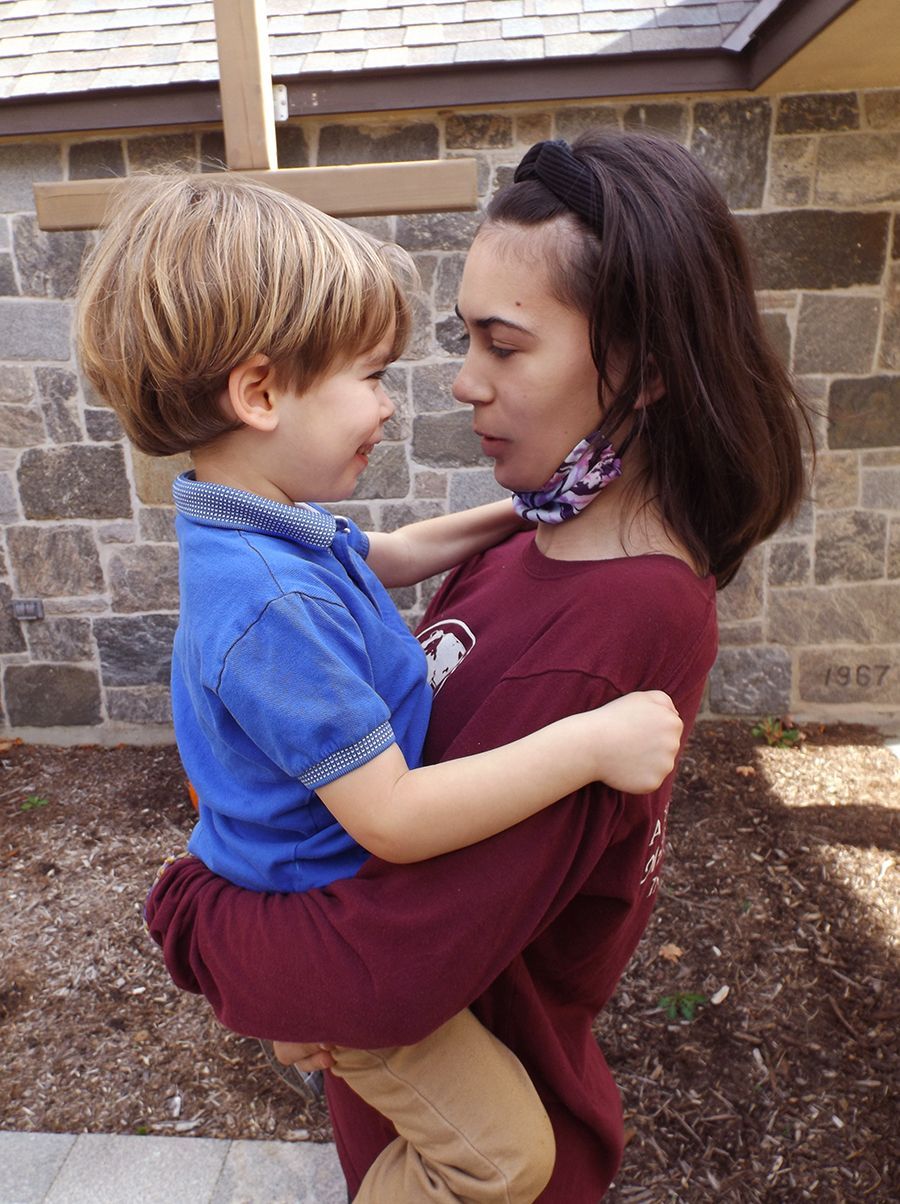 A girl holds a smiling little boy outdoors by a large wood cross near a stone wall while they gaze in profile at each other. 