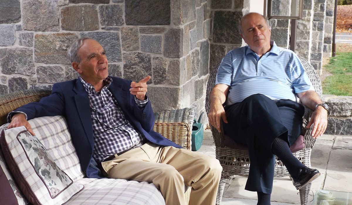 Two men sit on the church's stone porch, one gesturing while they talk.