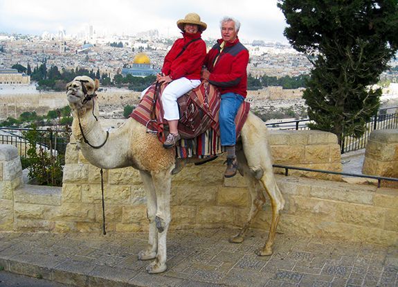 A husband & wife ride a camel together, on a stone terrace with a panoramic city view in the background