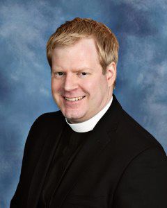 Smiling Pastor Charlie wears black clerical attire with white collar against a blue studio background