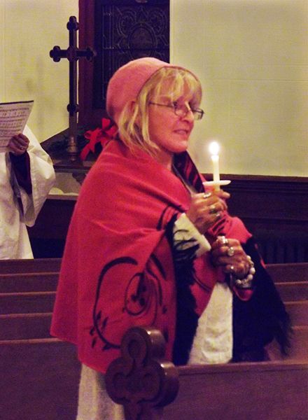 A woman in a pink hat & cloak stands in a pew holding a lit candle during Christmas Eve candlelight worship service