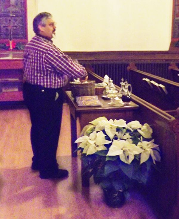 A male usher stands in the back of a church sanctuary at a table behind the pews, w/a white poinsettia plant nearby.