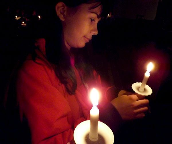 A girl in a red coat looks down reverently at two lit white candles she's holding  in a dark, candlelit church setting.