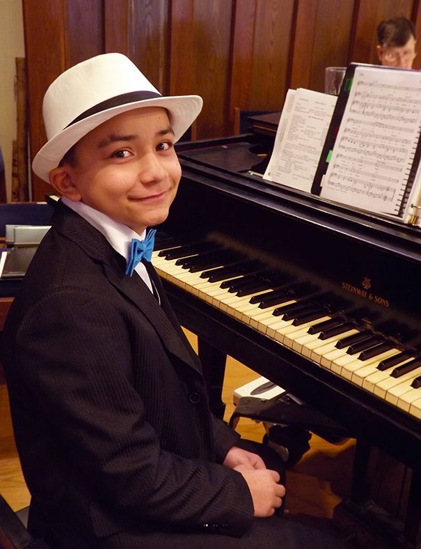 A young boy in a white hat & blue bow tie sits at a piano w/sheet music