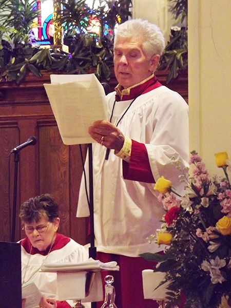 A man in a red gown and white choir robe singing a solo at a microphone on the altar during worship service.