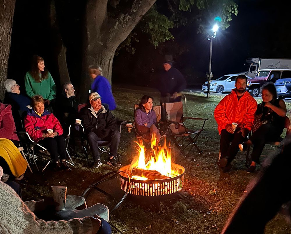 People gathered round a campfire at night in camping chairs under a large tree, near a parking lot with a bright light