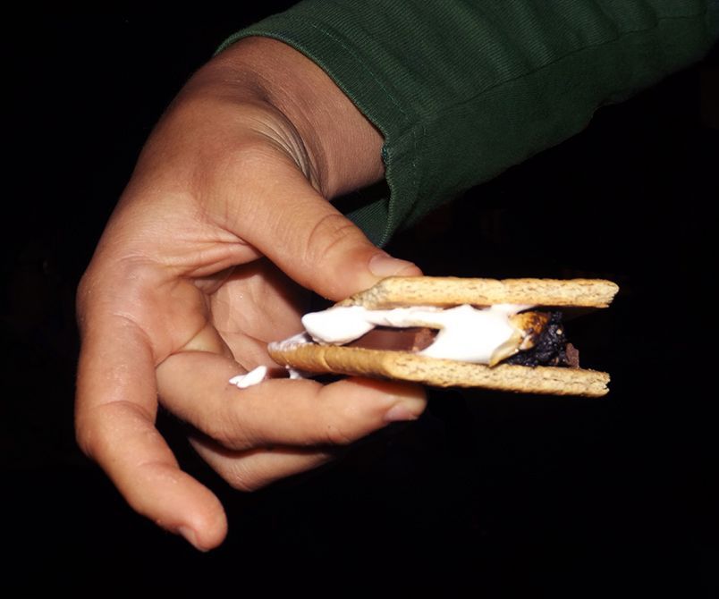 A hand holds a s’more treat with chocolate & melted marshmallow on a graham cracker, against the dark nighttime background.