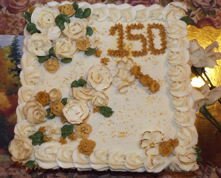 An elaborately decorative white cake with gold roses and the number “150” in gold icing on top waits to be sliced