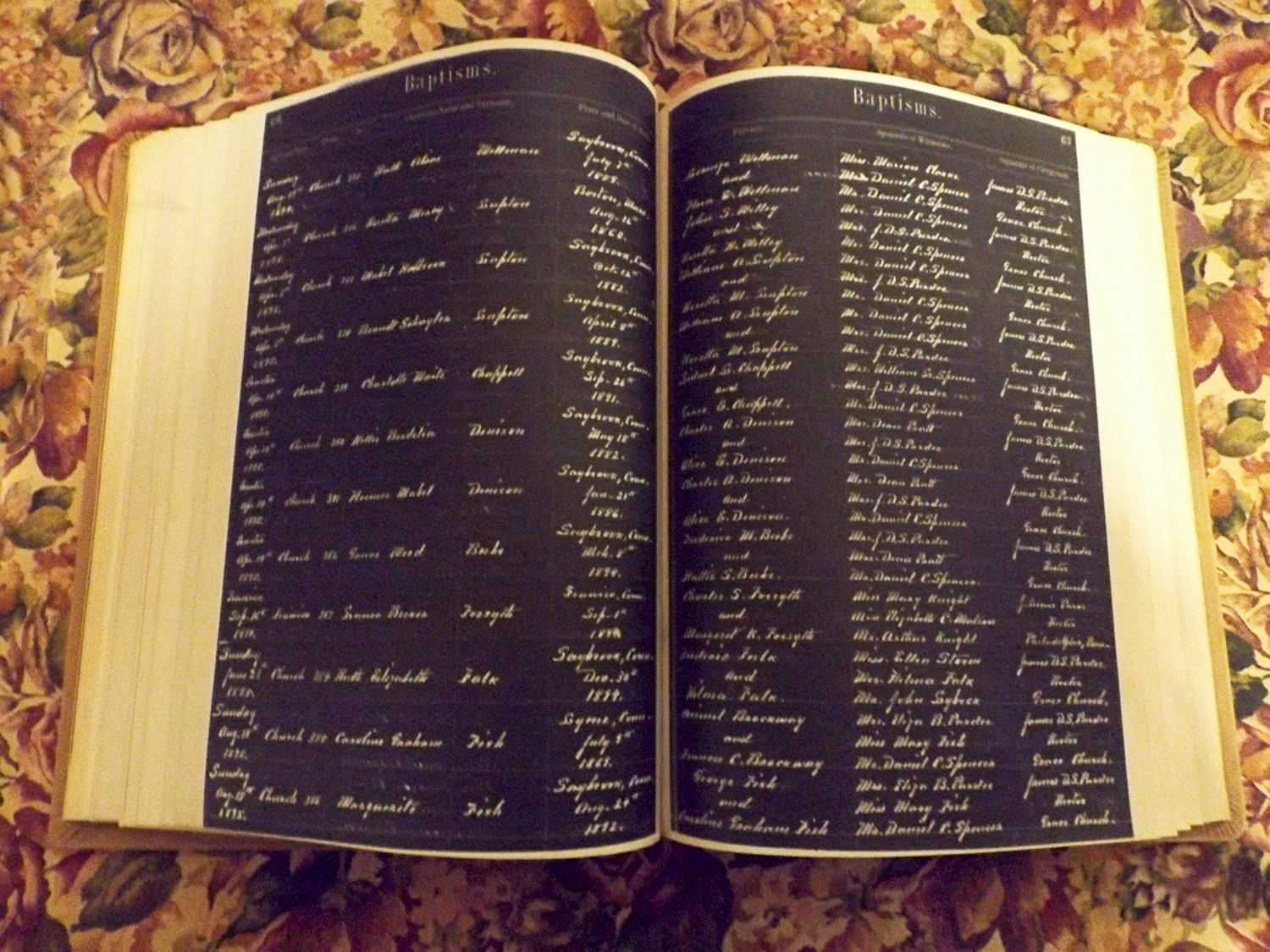 A hardbound historical archive handwritten on a black background sits open to a BAPTISMS page atop a floral fabric tablecloth