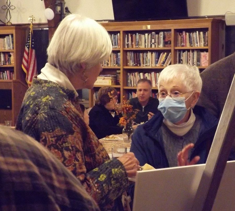 Two women (one masked) converse during a special exhibit in the Parish Hall with a placard nearby & bookshelves in the rear