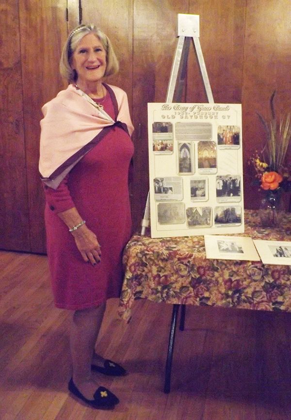 A welcoming woman in a red dress stands beside a display board at a table in the Parish Hall