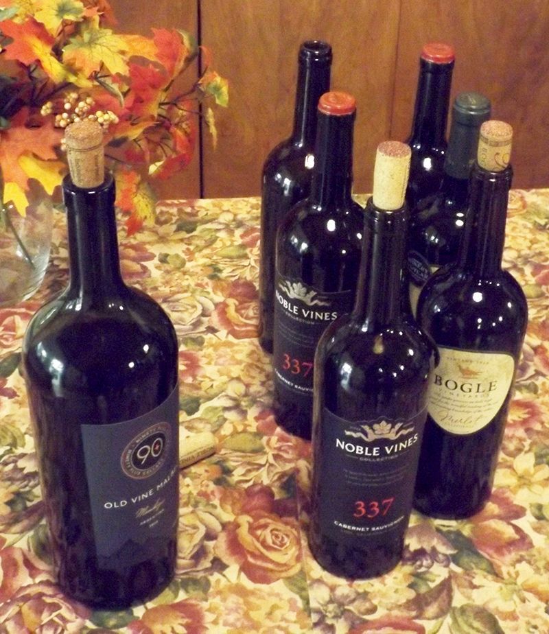 Seven wine bottles rest on a patterned tablecloth, near a vase with autumn leaves
