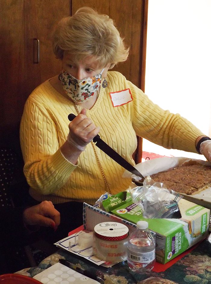 A woman in a yellow sweater w/name-tag & floral mask wields a large serving-knife at the baked goods table