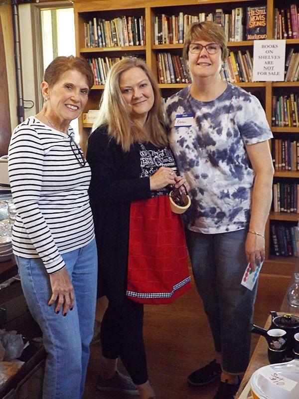 Three smiling women stand in front of a bookshelf in the Parish Hall, preparing for the annual Tag Sale.