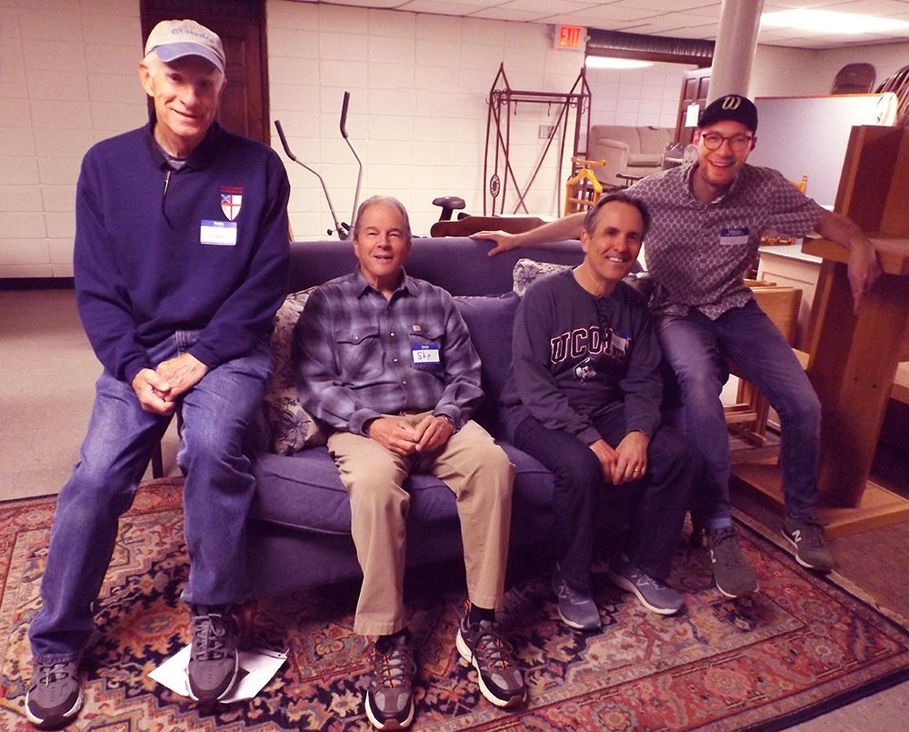 A group of four men take a rest from work, on a couch in the basement, atop a patterned rug.