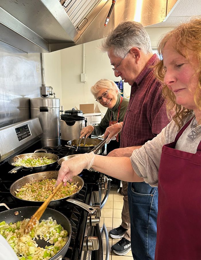 A man & 2 women cook together at a stovetop under a large  range hood, in a bright kitchen, stirring food in skillets.