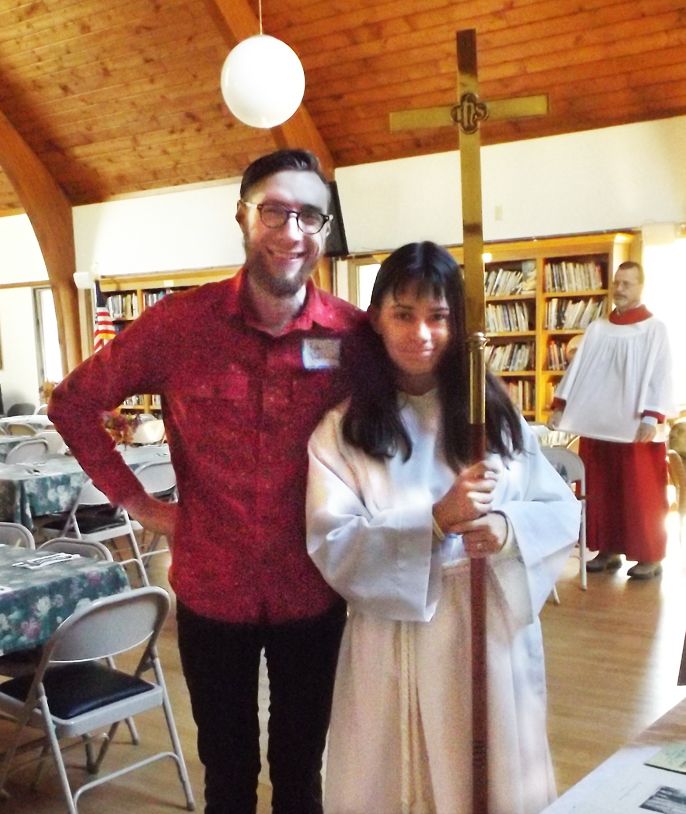 A male parishioner stands with a female acolyte in a white robe holding a cross staff, with bookshelves behind them 